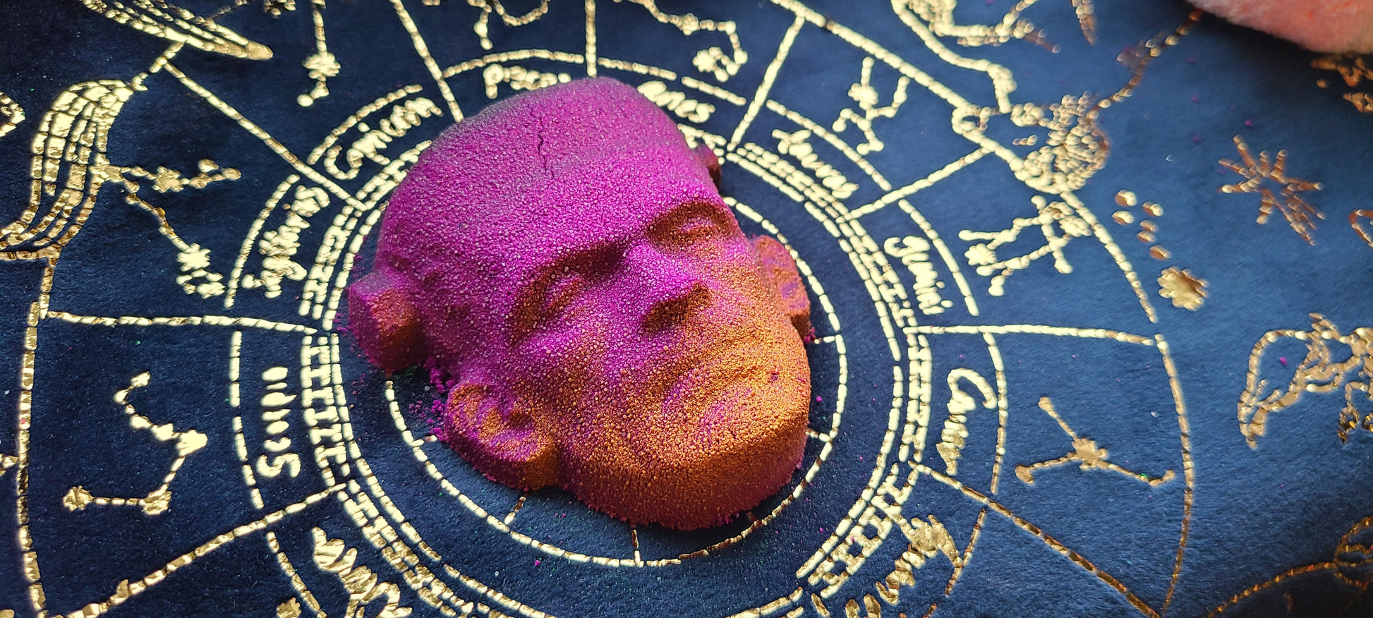 Dark pink Frankenstein face bath bomb with golden highlights on a dark blue and gold astrology background