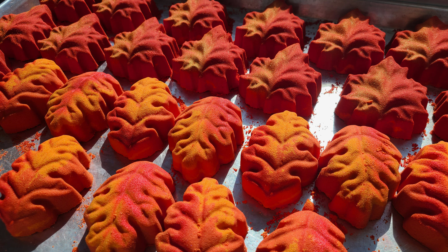Maple leaf bath bomb that is red and gold sitting on a tray with Oak leaf bath bombs that are yellow and orange with red glitter